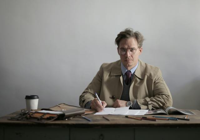 mężczyzna w prochowcu siedzi przy biurku z dokumentami